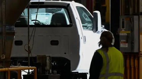 Bloomberg via Getty Images A Ford truck on a production line, passing through inspection checks, at a plant in Ohio. A female worker wearing a high-vis jacket looks on.
