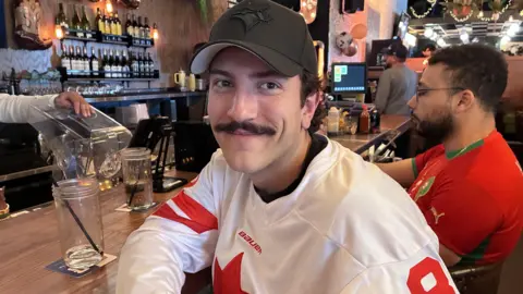Matt Lorincz, with a moustache, wears a black Blue Jays baseball cap and a white and red Canada jersey. He is smiling and resting one arm on a bar counter.