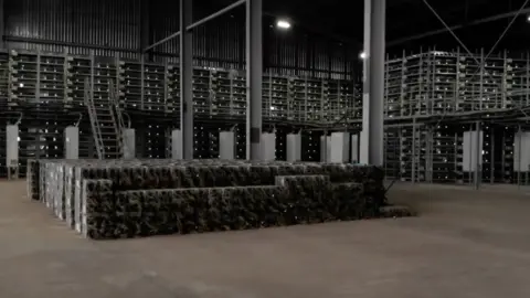 Black racks of powerful computers in a warehouse. They are surrounded by scaffolding and have white dotted lights on them.