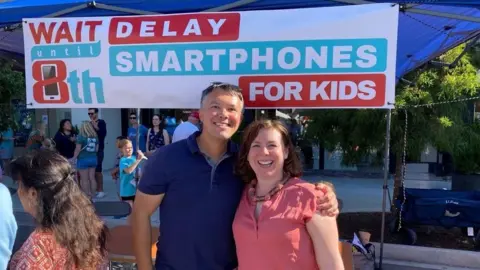 Handout Mark SooHoo advocates for children to delay their use of smartphones. He stands with a woman in front of a banner that says Wait Until 8th