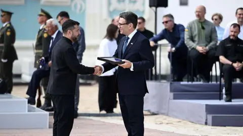 AFP via Getty Images Zelensky and Burnham shake hands and smile as the latter holds a frame