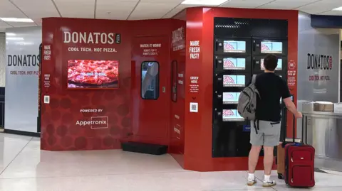 Appetronix The dark red Donatos fresh pizza vending machine is about the size of a van. A customer is looking at its screen.