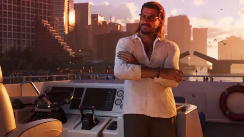 Rockstar Games A male character from GTA 6 in an open-collared shirt with slicked-back hair and sunglasses leans, arms folded, against the controls of a yacht as it coasts down a waterway flanked by skyscrapers. A road bridge opens in the distance as orange dawn light reflects of the glass fronts of the tall buildings. The computer-generated image is convincingly lifelike.