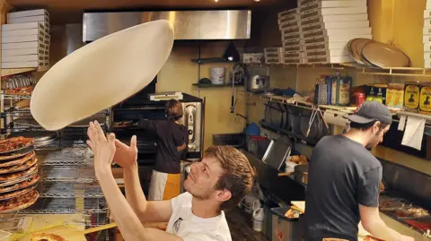 Portland Press Herald via Getty Images A chef tosses the base of a pizza into the air, behind him other kitchen staff work on pizzas.