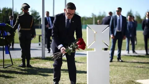 AFP via Getty Images Burnham bends down holding a bunch of red roses in front of a white gravestone as people stand behind him
