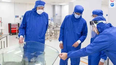 Saint-Gobain Four workers in blue overalls inspect an aircraft window. They are wearing masks and hoods.