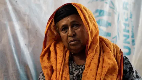 Liam Weir/BBC An elderly woman wearing a headscarf as she sits on the floor in the al-Monig displacement camp