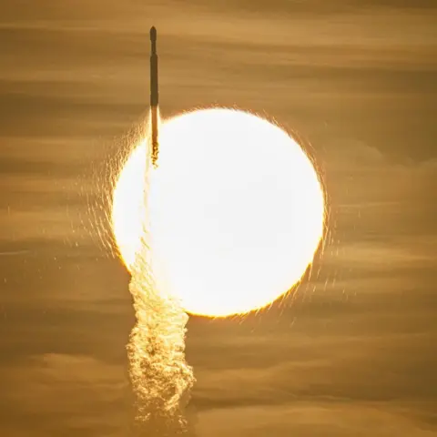 NASA/John Kraus A view of a rocket launching off to space in front of the blaring yellow sun and yellow-tinted sky