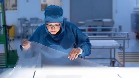 Saint-Gobain A Saint-Gobain worker in a darkened room works on a clear sheet of acrylic.