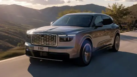Bentley An aluminium-coloured Bentley SUV is driven on a road with hills and countryside behind it.
